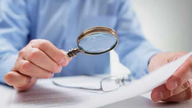 A person examining a document closely with a magnifying glass, suggesting detailed scrutiny or the due diligence process.