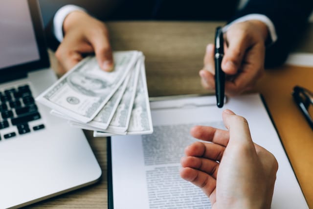 A person in a business suit offering a stack of US dollar bills to someone reaching out, with a laptop and documents on the desk, indicating a financial transaction or deal in progress.
