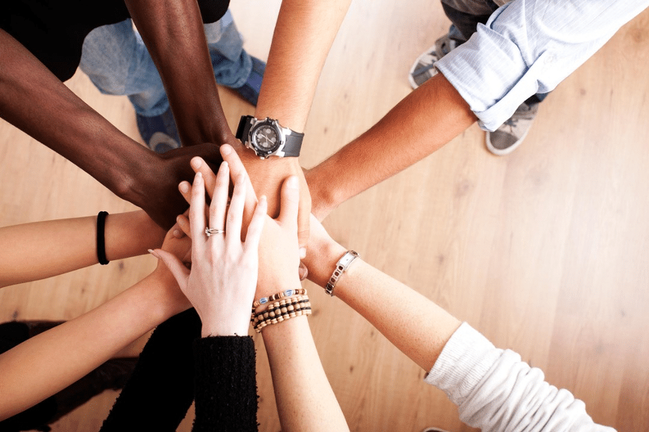 A top-down view of a group of people standing in a circle, placing one hand each in the center, symbolizing teamwork, unity, preparation and collaboration.