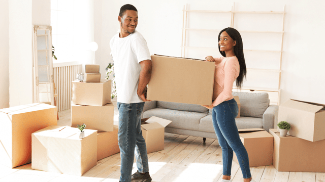 Smiling man and woman carrying a large cardboard box in a bright, airy room filled with packed boxes, indicating they are moving into a new home
