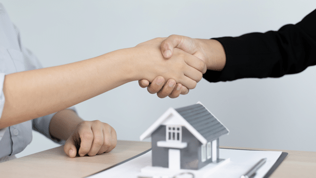 Two individuals shaking hands over a desk with a small model house on it, highlighting the role of women in real estate.