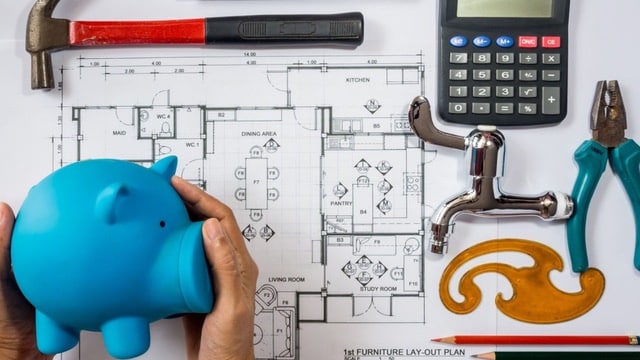 Blueprint of a house layout with a piggy bank, calculator, and tools representing budgeting for house flipping costs.