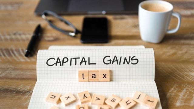 Notebook with 'CAPITAL GAINS tax' spelled out in letter tiles, representing 2024 Capital Gains Tax Changes, alongside a coffee cup, pen, and digital tablet.