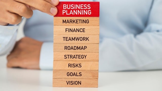 A stack of wooden blocks with business planning concepts written on them, including 'VISION,' 'GOALS,' 'RISKS,' 'STRATEGY,' 'ROADMAP,' 'TEAMWORK,' 'FINANCE,' and 'MARKETING' at the top in red. A person in a business shirt is in the background, implying a focus on structured planning in a professional setting.