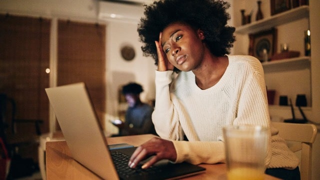 A focused woman, embodying resilience for real estate investors, works intently on her laptop in a cozy evening home setting, with a person in the background.