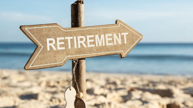  a rustic wooden sign in the shape of an arrow, labeled "RETIREMENT", pointing to the right. The sign is attached to a wooden post and is set against a serene beach backdrop, with clear blue skies and calm seas. 