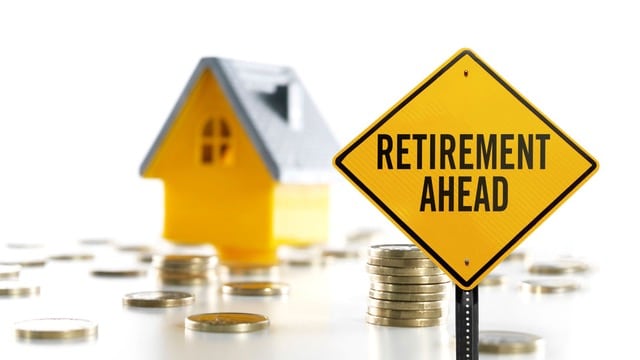 The image features a yellow road sign with the words "RETIREMENT AHEAD" prominently displayed. In the background, there are two miniature houses, one yellow and one blue, symbolizing property or real estate investment. Scattered around the houses are numerous coins, representing financial savings or investments leading up to retirement.