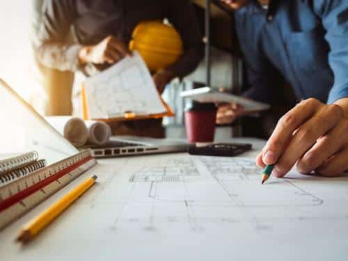 Close-up of architectural plans and drawings on a desk, with engineers reviewing documents and discussing the land development process in the background.